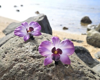 May include: Two purple orchid hair clips on a gray rock. The orchids have a white center and a yellow center.