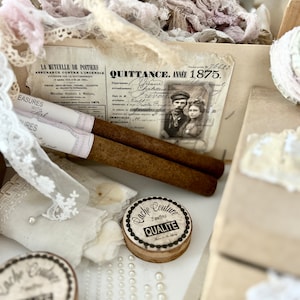 Könnte beinhalten: Eine antike Holzkiste gefüllt mit Spitze, Stoffresten und einer kleinen runden Dose mit der Aufschrift "Cache Couture 2 Meter Qualite". Ein Stück Papier mit dem Text "QUITTANCE. ANNÉE 1,875." ist sichtbar.