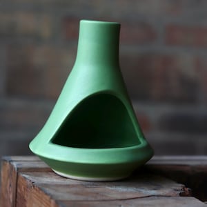 May include: A matte light green ceramic oil burner with a conical shape and a wide, arched opening at the base. The object features a smooth, uniform finish and a flat, circular bottom, designed with a hollow interior for holding a heat source.