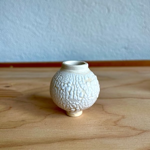 May include: A small, white ceramic vase with a textured surface. The vase has a round body and a small, round base.