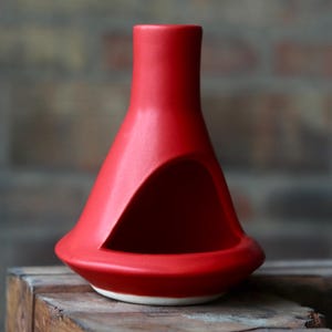 May include: A bright red ceramic oil burner with a conical shape and a wide, circular base. The object features a hollowed-out triangular opening on one side for access to the interior, with a smooth matte finish and a plain white underside.