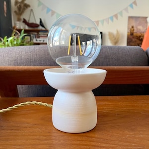 May include: A small, white ceramic table lamp with a clear, round lightbulb. The lamp has a unique, hourglass-shaped base and a gold-colored cord. The lightbulb has a visible filament. The lamp is on a wooden table.