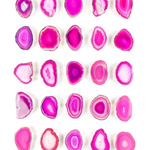 May include: A collection of twenty-five polished pink agate slices, each with a gold-coloured edge. The slices display a variety of shapes and internal patterns, including concentric rings and translucent sections. The background is plain white, accentuating the gemstones' colours and textures.
