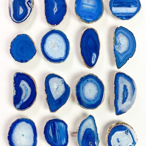 May include: A collection of blue agate slices with gold-plated edges. The polished stones vary in shape and size, showcasing concentric patterns of white and varying shades of blue. These decorative pieces are ideal for home decor.