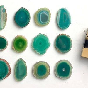 May include: Twelve green agate coasters with a variety of patterns and shapes. The coasters are arranged in three rows of four. A brown gift box with a black bow is in the top right corner of the image.