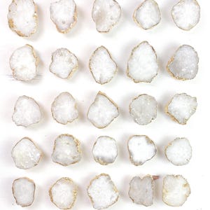 Op de afbeelding: Een verzameling van vierentwintig witte geode kastknoppen met goudkleurige randen. De knoppen zijn onregelmatig van vorm en gerangschikt in een rasterpatroon tegen een witte achtergrond. De geode knoppen hebben een kristalachtige textuur.