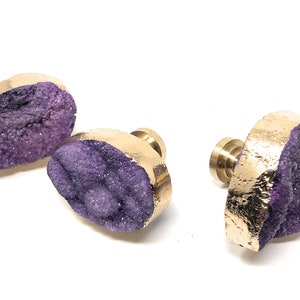May include: Three purple agate crystal drawer pulls with gold accents. The crystal is a natural geode with a rough, textured surface. The pulls are mounted on a brass base.
