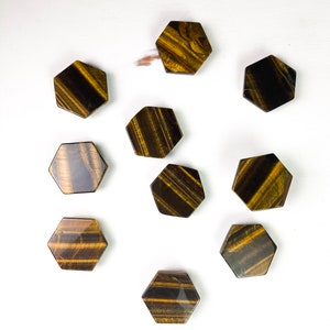 May include: Ten hexagonal tiger's eye stones, each with a different pattern of brown and gold stripes, arranged in a circle on a white surface.