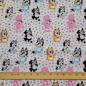 May include: White fabric with a repeating pattern of cartoon dogs in various colors, including blue, orange, and black. Pink poodles and colorful confetti-like shapes are also scattered across the fabric. A wooden ruler is placed at the bottom.