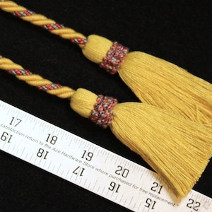 May include: Two yellow tassels with twisted gold and red cords. The tassels have a decorative band of red, blue, and purple threads. The image includes a ruler with text, indicating the tassels' potential use in crafts or decor.