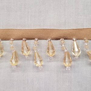 May include: A tan fabric trim featuring a row of dangling, faceted, teardrop-shaped beads in a translucent golden yellow color. The beads are attached to the ribbon by small, clear, round spacer beads, creating a decorative fringe border.