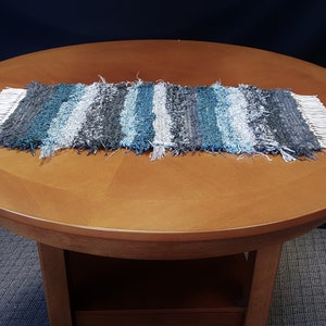 May include: A woven table runner with a blue, gray, and white striped pattern. The runner has a fringe on both ends.