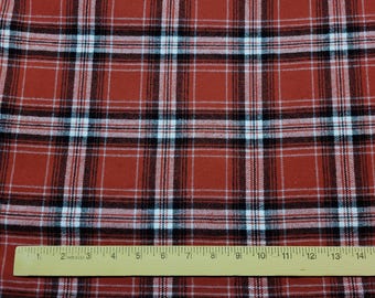 May include: Close-up of a red, black, and white plaid fabric. The fabric has a classic tartan pattern with intersecting lines forming squares. A wooden ruler with inch markings is at the bottom, measuring up to 15 inches.