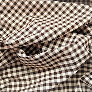 May include: Brown and white gingham fabric with a checkered pattern. The fabric is folded and draped, showing the texture and weave.