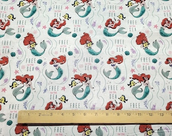 May include: White fabric with a repeating pattern of Ariel the Little Mermaid. Ariel is depicted as a mermaid with red hair, a green tail, and a seashell bra. She is holding a seashell and a yellow fish. The fabric also features the text "Free as the Sea" in pink and purple.