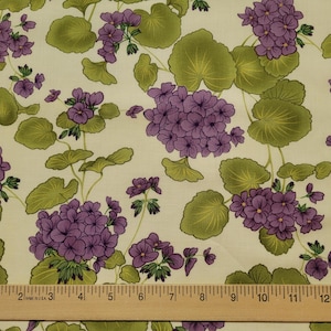 May include: Cream-colored fabric with a repeating pattern of purple geranium flowers and green leaves. The flowers are clustered, and the leaves are large and rounded. A wooden ruler is at the bottom.