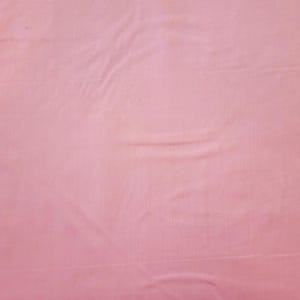 May include: A solid pink fabric with visible folds and creases. The material has a soft, matte appearance and a subtle sheen. The color is a light, muted shade of pink, with variations in tone due to the fabric's texture.