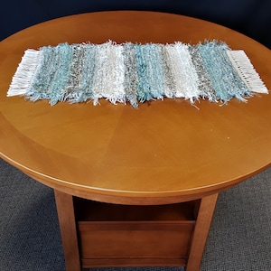 May include: A woven table runner with a fringe on each end. The runner is made of white, blue, and gray yarn and is laid on a round wooden table.