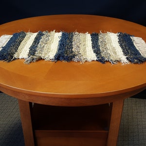 May include: A woven table runner with blue, white, and brown stripes. The runner has a fringed edge and is made of a soft, textured material.
