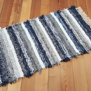 May include: A striped rug with a fringe border. The rug is made of recycled denim and features shades of blue, white, and gray.