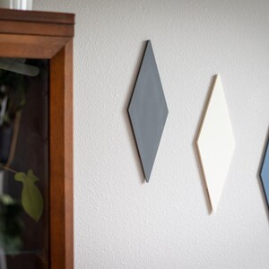 Puede incluir: Tres decoraciones de pared en forma de diamante en gris, blanco y azul est&aacute;n montadas en una pared blanca texturizada. Las formas geom&eacute;tricas est&aacute;n dispuestas verticalmente, agregando un toque moderno. Un mueble con marco de madera es visible a la izquierda.