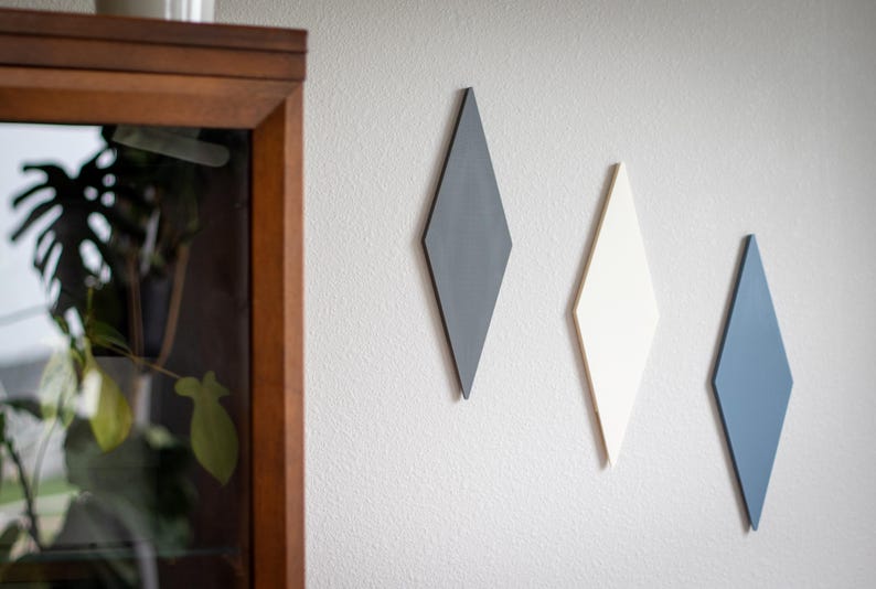 Puede incluir: Tres decoraciones de pared en forma de diamante en gris, blanco y azul est&aacute;n montadas en una pared blanca texturizada. Las formas geom&eacute;tricas est&aacute;n dispuestas verticalmente, agregando un toque moderno. Un mueble con marco de madera es visible a la izquierda.