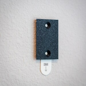 Puede incluir: Una tira rectangular gris oscuro para colgar cuadros con dos agujeros y una pesta&ntilde;a adhesiva blanca con el logotipo "3M" y una flecha hacia abajo. La tira est&aacute; montada en una pared texturizada de color blanquecino.