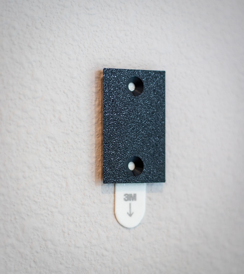 Puede incluir: Una tira rectangular gris oscuro para colgar cuadros con dos agujeros y una pesta&ntilde;a adhesiva blanca con el logotipo "3M" y una flecha hacia abajo. La tira est&aacute; montada en una pared texturizada de color blanquecino.