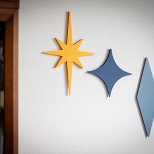 Puede incluir: Tres piezas de decoraci&oacute;n de pared modernas de mediados de siglo en amarillo, azul y azul claro. Las formas incluyen una estrella, un diamante y un rombo, todos montados en una pared blanca. La decoraci&oacute;n a&ntilde;ade un toque retro a cualquier habitaci&oacute;n.