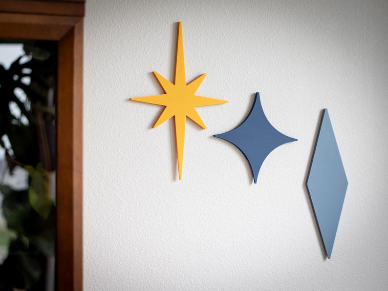 Puede incluir: Tres piezas de decoraci&oacute;n de pared modernas de mediados de siglo en amarillo, azul y azul claro. Las formas incluyen una estrella, un diamante y un rombo, todos montados en una pared blanca. La decoraci&oacute;n a&ntilde;ade un toque retro a cualquier habitaci&oacute;n.