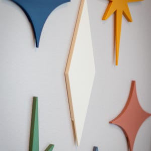 Puede incluir: Decoraci&oacute;n mural geom&eacute;trica y colorida, compuesta por estrellas y diamantes. Las piezas est&aacute;n pintadas en tonos de azul, naranja, verde y coral, creando una est&eacute;tica moderna de mediados de siglo. El arte mural est&aacute; montado en una pared gris claro.
