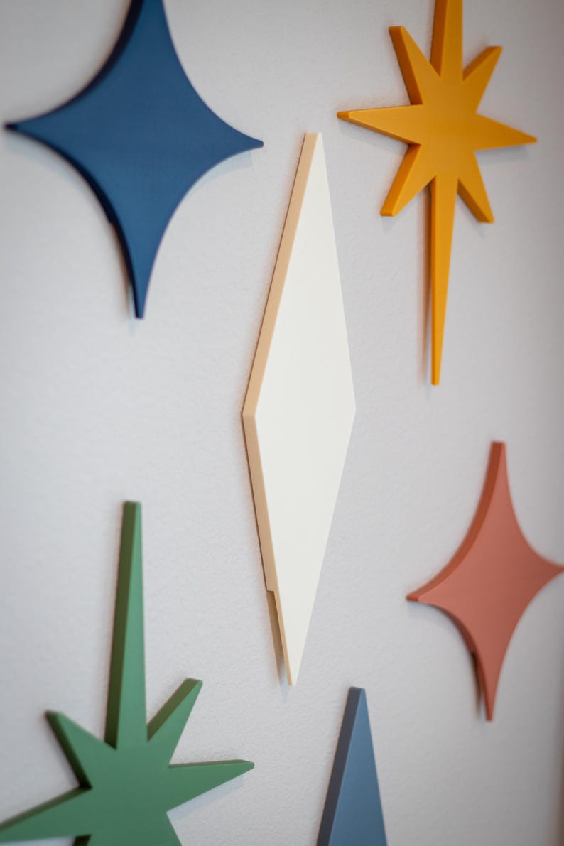 Puede incluir: Decoraci&oacute;n mural geom&eacute;trica y colorida, compuesta por estrellas y diamantes. Las piezas est&aacute;n pintadas en tonos de azul, naranja, verde y coral, creando una est&eacute;tica moderna de mediados de siglo. El arte mural est&aacute; montado en una pared gris claro.