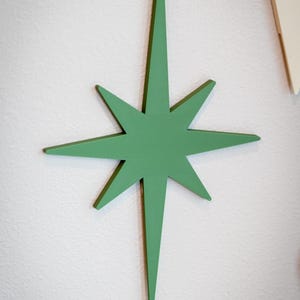 Puede incluir: Decoraci&oacute;n de pared en forma de estrella verde de ocho puntas. La estrella est&aacute; pintada de verde liso y est&aacute; montada en una pared blanca. Esta pieza decorativa a&ntilde;ade un toque moderno de mediados de siglo.