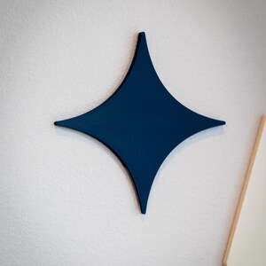 Puede incluir: Decoraci&oacute;n de pared en forma de estrella de cuatro puntas, de color azul marino. La estrella est&aacute; montada en una pared blanca con superficie texturizada. El dise&ntilde;o de la estrella es simple y moderno, adecuado para varios estilos de interior.