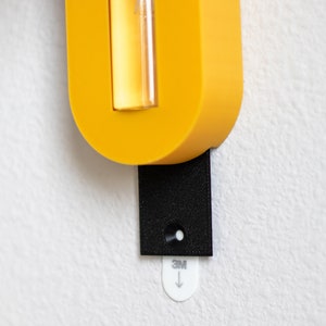 Puede incluir: Un soporte de pared amarillo con un vial de vidrio transparente y una tira adhesiva negra con el texto "3M" y una flecha hacia abajo.