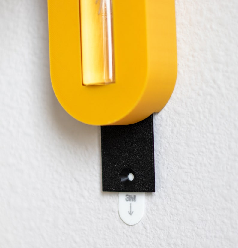 Puede incluir: Un soporte de pared amarillo con un vial de vidrio transparente y una tira adhesiva negra con el texto "3M" y una flecha hacia abajo.