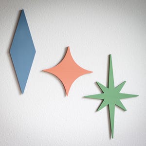 Puede incluir: Tres piezas de decoraci&oacute;n de pared de madera con formas geom&eacute;tricas. Un rombo azul, una estrella de coral y una estrella verde est&aacute;n montadas en una pared blanca. Estas decoraciones de estilo moderno de mediados de siglo a&ntilde;aden un toque de color y un toque retro.