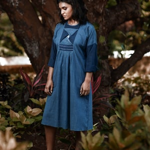 Könnte beinhalten: Eine Frau trägt ein jeansblaues Kleid und eine passende Jacke. Das Kleid hat einen plissierten Rock und ein eng anliegendes Oberteil. Die Jacke hat Dreiviertelärmel. Der Hintergrund zeigt einen großen Baum und üppiges Grün.