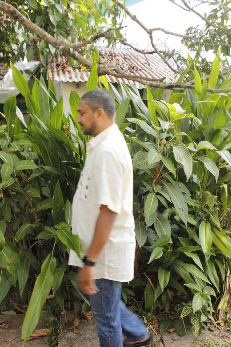 May include: A person wearing a light-colored short-sleeved shirt and blue jeans walks past lush green foliage. The background includes a glimpse of a building with a white roof and a tree branch.
