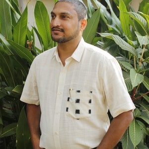 May include: Cream-colored short-sleeved shirt with a pocket featuring a black and white checkered pattern. The shirt is buttoned and worn with blue jeans. A pen is visible in the pocket. The person is wearing a black watch.