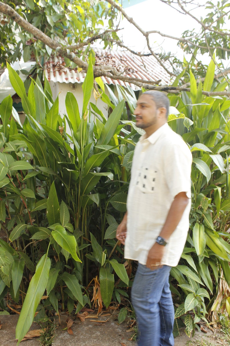 May include: A person wearing a light yellow short-sleeved shirt with a black square pattern on the pocket and blue jeans, walking past lush green plants. A watch is visible on the person's wrist. The background includes a white roof and tree branches.