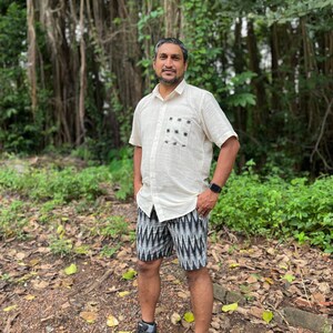 May include: A man wearing a cream-colored short-sleeved shirt with a black and white patterned pocket and black and white patterned shorts. He is wearing gray sneakers and a black smartwatch. The background features lush green foliage.