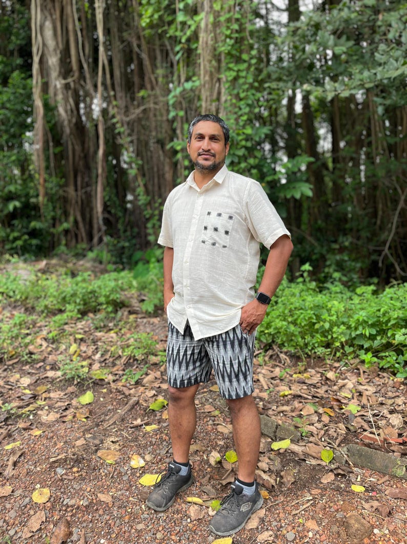 May include: A man wearing a cream-colored short-sleeved shirt with a black and white patterned pocket and black and white patterned shorts. He is wearing gray sneakers and a black smartwatch. The background features lush green foliage.