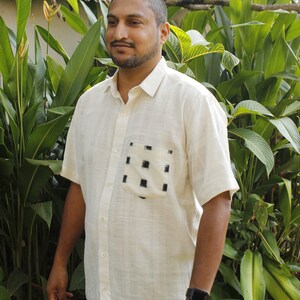May include: Off-white short-sleeved shirt with a button-down front and a chest pocket featuring a black square pattern. The shirt is made of a textured fabric and is worn with blue jeans. A black smartwatch is visible on the wrist.