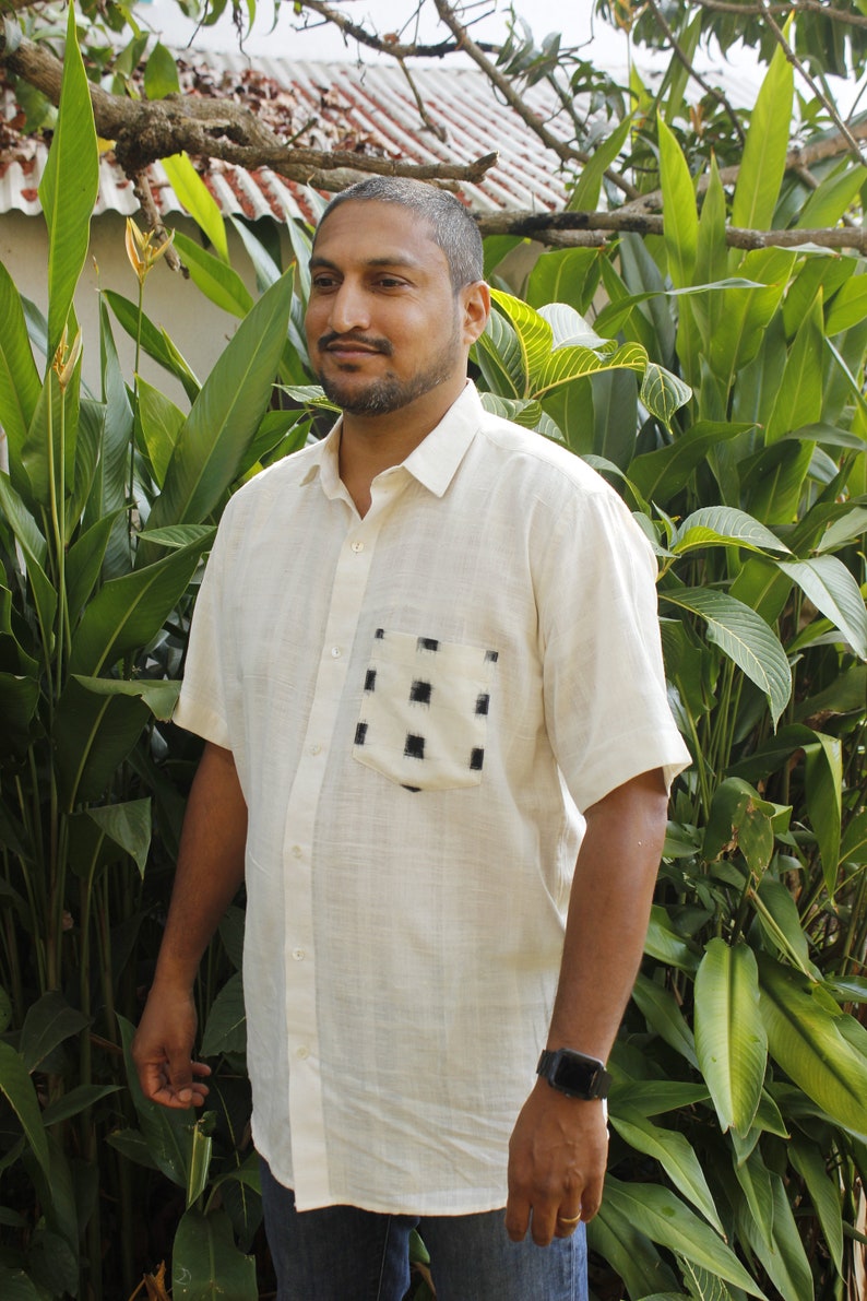 May include: Off-white short-sleeved shirt with a button-down front and a chest pocket featuring a black square pattern. The shirt is made of a textured fabric and is worn with blue jeans. A black smartwatch is visible on the wrist.