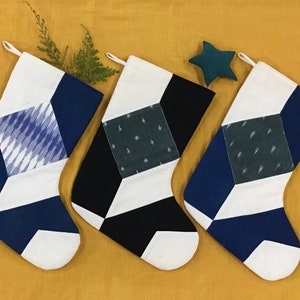 以下が含まれることがあります： ネイビー、白、黒の幾何学模様のクリスマスストッキング3つ。1つは青と白のイカット柄、他は水玉模様です。緑の星と緑の小枝も写っています。