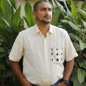 May include: A cream-colored short-sleeved shirt with a black and white checkered pocket. The shirt is paired with blue jeans. The person is standing in front of green foliage.