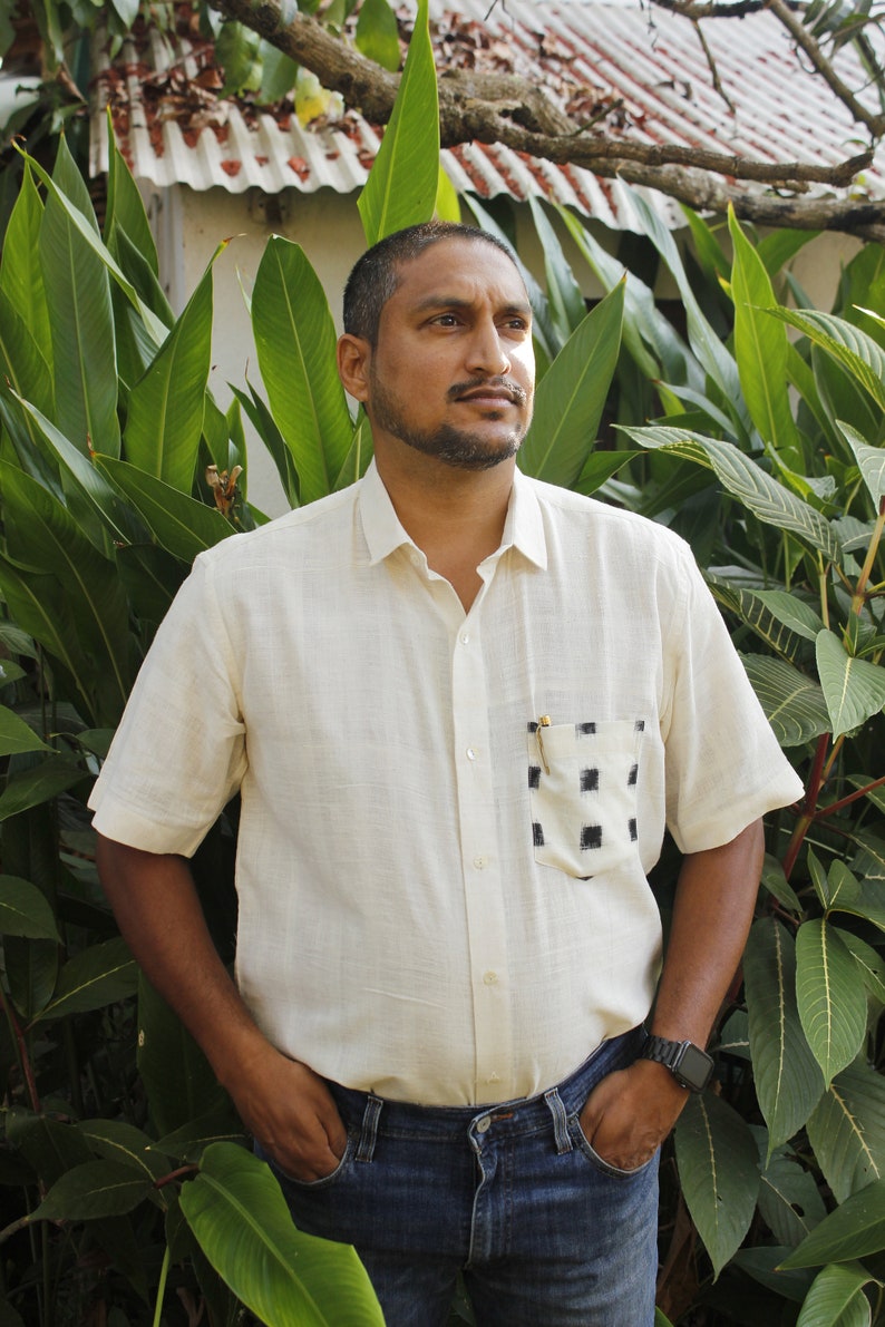 May include: A cream-colored short-sleeved shirt with a black and white checkered pocket. The shirt is paired with blue jeans. The person is standing in front of green foliage.
