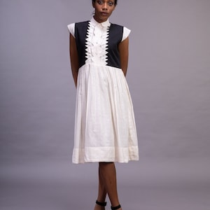 May include: A sleeveless white dress with a black bodice and a pleated skirt. The bodice features a geometric design. The model is wearing black high-heeled sandals. The dress has a collared neckline.