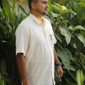 May include: A light beige short-sleeved button-down shirt with a pocket, paired with blue jeans. The shirt is worn by a person standing in front of lush green foliage. A watch is visible on the person's wrist.
